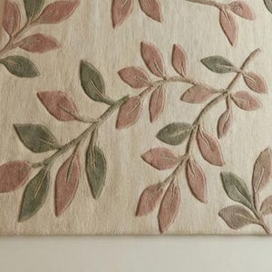 May include: Close-up of a beige rug with an embossed leaf design. The leaves are in shades of pink and green, arranged on branches across the surface. The rug has a textured appearance, suggesting a soft, comfortable material.