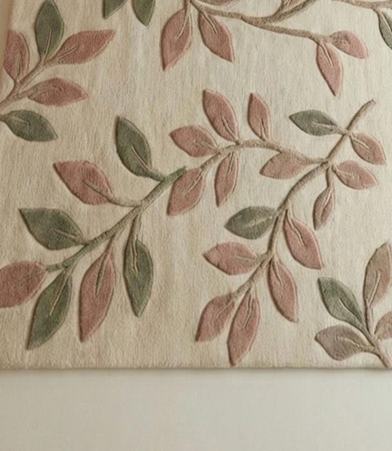 May include: Close-up of a beige rug with an embossed leaf design. The leaves are in shades of pink and green, arranged on branches across the surface. The rug has a textured appearance, suggesting a soft, comfortable material.