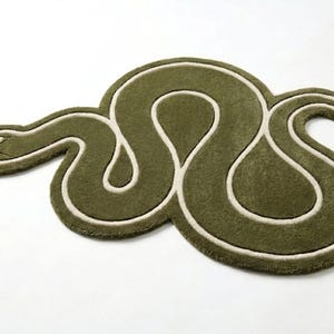 Puede incluir: Una alfombra de mechones con forma de serpiente enroscada. La alfombra presenta un color verde oliva intenso con un contorno en color crema que sigue el cuerpo sinuoso de la serpiente, incluyendo una cabeza detallada con ojo y boca.