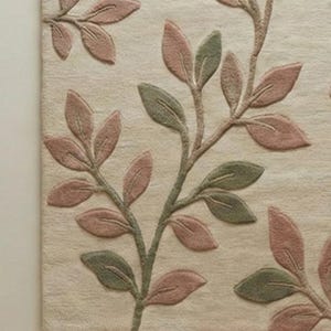 May include: Beige rug with a raised leaf pattern in shades of pink and green. The leaves are arranged on branches that extend across the rug's surface. The rug has a soft, textured appearance, suitable for a living room or bedroom.
