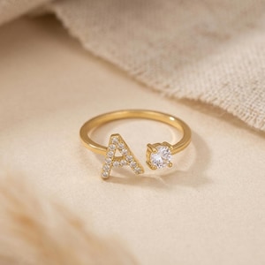 May include: Gold-colored ring featuring the letter "A" encrusted with small, clear gemstones. A single, larger clear gemstone is set opposite the letter. The ring is displayed on a neutral-toned surface.