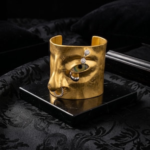 Op de afbeelding: Een goudkleurige manchetarmband met een gezichtsontwerp. De armband heeft een oog met een groene iris, een neus en een mond. Het oog heeft een diamant erboven en eronder. De neus heeft een diamanten ring. De armband ligt op een zwart oppervlak.