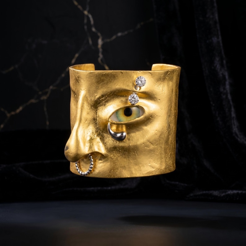 Op de afbeelding: Een goudkleurige manchetarmband in de vorm van een gezicht, met een oog met een groene iris en een zilveren traan. De armband heeft twee diamantaccenten boven het oog en een neusring. De manchet is geplaatst tegen een donkere achtergrond.