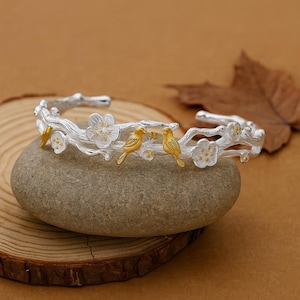 Può includere: Bracciale a polsino color argento con un design a ramo, dettagli di fiori bianchi e due uccelli color oro. Il bracciale è appoggiato su una pietra grigia, mostrando i suoi dettagli e la sua lavorazione.
