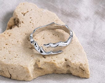 May include: A silver-coloured, open-ended ring with a textured, organic design. The ring features an irregular, wave-like pattern around the band. The ring is displayed on a light-coloured, textured stone.