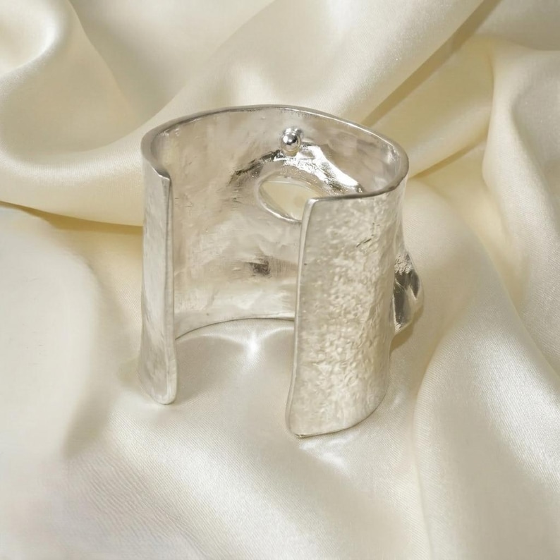 Op de afbeelding: Een zilverkleurige manchetarmband met een getextureerd oppervlak. Het open ontwerp heeft een gehamerd metaaleffect en een klein decoratief element aan de bovenkant. De armband wordt gepresenteerd op een witte, zijdeachtige stof.