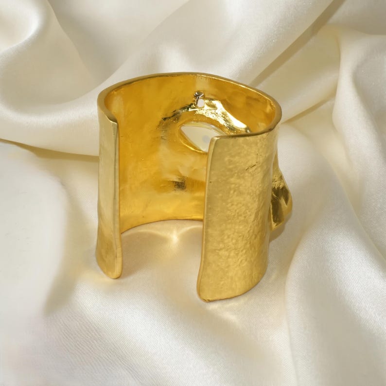 Op de afbeelding: Een goudkleurige manchetarmband met een getextureerd oppervlak. Het open ontwerp heeft een afgeronde vorm met een centrale uitsparing. De armband wordt getoond op een witte, zijdeachtige stof.