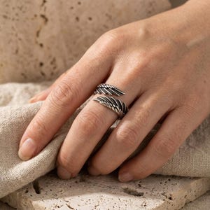 May include: A silver-toned metal ring featuring an open-ended design shaped like two overlapping bird wings. The surface of the ring has a textured, oxidized finish that highlights the detailed feather carvings along the length of each wing.