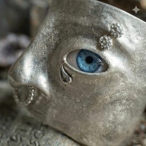 Op de afbeelding: Een zilveren masker met een gedetailleerd gezicht, met een helder blauw oog met een traandetail. Het masker heeft een getextureerd oppervlak en kleine decoratieve elementen bij het oog en de mond.