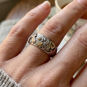May include: A wide band ring featuring a mixed metal design with silver, gold, and copper tones. The surface is decorated with embossed floral motifs, including a central daisy, and stylised dragonfly figures integrated into the metalwork.