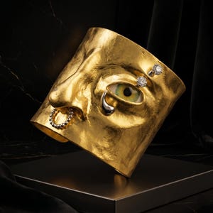 Op de afbeelding: Een goudkleurig gezichtsmasker met een neusring, een traan en twee diamanten oorbellen. Het masker heeft een groen en geel oog. Het masker wordt getoond op een zwart oppervlak tegen een zwarte achtergrond.