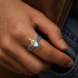 May include: A silver ring with a detailed eagle design. The eagle's head is gold-colored, and the wings are spread wide. The ring is worn on a finger, showcasing the intricate craftsmanship and the bird's majestic pose.