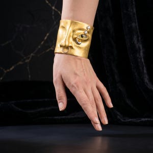 Op de afbeelding: Een goudkleurige manchetarmband met een gebeeldhouwd gezichtsontwerp. De armband heeft een neus, een oog met een edelsteen en een neusring. Twee extra edelstenen zijn aan de zijkant gezet. De sieraden worden om de pols gedragen.