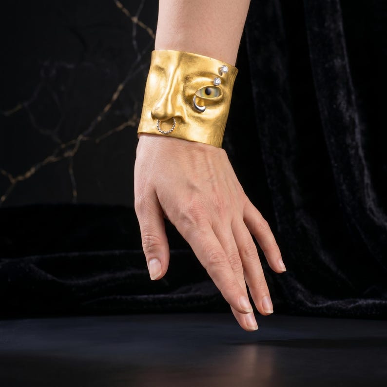 Op de afbeelding: Een goudkleurige manchetarmband met een gebeeldhouwd gezichtsontwerp. De armband heeft een neus, een oog met een edelsteen en een neusring. Twee extra edelstenen zijn aan de zijkant gezet. De sieraden worden om de pols gedragen.