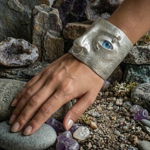 Op de afbeelding: Een zilveren manchetarmband met een gezichtsontwerp, met een blauw oog, een neus en een kleine oorbel. De armband wordt om de pols gedragen, met de hand rustend op een bed van rotsen en kristallen. Een uniek sieraad.