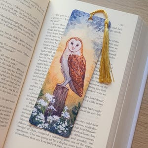 May include: Watercolour bookmark depicting a barn owl on a wooden post, with white flowers. The bookmark has a gold tassel and is set against the pages of a book.