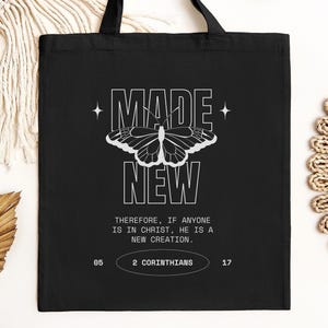 May include: Black tote bag with the words "MADE NEW" in white block letters, a butterfly graphic, and a Bible verse. The bag has black handles and is made of a sturdy fabric. The text "2 Corinthians 17" is also visible.