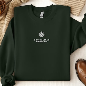 May include: Dark green sweatshirt with the text "O COME, LET US ADORE HIM" and a compass rose design. The sweatshirt is displayed on a beige knitted blanket, with brown leather shoes and a beige beanie in the background.