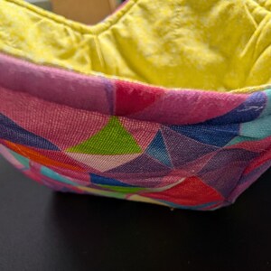 May include: A colorful bowl cozy with a geometric pattern in pink, blue, green, and red. The interior is a bright yellow. The bowl cozy is designed to keep food warm and is made of fabric.