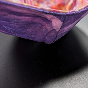 May include: A fabric bowl with a purple, marbled exterior and a colorful floral interior. The bowl is square-shaped with a folded design, showcasing a blend of purple, pink, and white hues. The interior features a vibrant mix of pink, orange, and yellow.