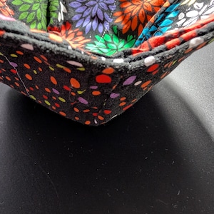 May include: A handmade bowl cozy with a black exterior covered in colorful polka dots. The interior features a floral pattern with orange, purple, green, and yellow flowers on a black background. The bowl cozy is designed to keep food warm.