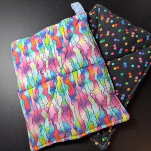 May include: Two quilted pot holders. One has a colorful wavy pattern in shades of pink, blue, yellow, and green. The other is black with a pattern of colorful polka dots. Both have a loop for hanging.