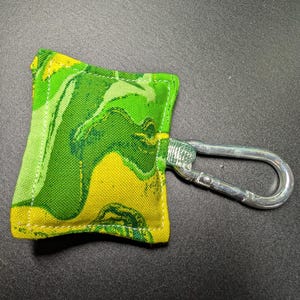 May include: A small, square fabric item with a green and yellow marbled pattern, attached to a silver-colored carabiner clip. The fabric has a stitched border. The item is likely a small pouch or accessory.