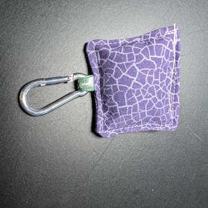 May include: A small, square, purple fabric item with a white, cracked pattern, attached to a silver carabiner clip. The fabric is stitched with white thread. The clip is attached to a green and white fabric loop.