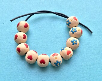 Puede incluir: Una cadena de cuentas de color crema con corazones rojos y flores azules pintadas sobre ellas. Las cuentas están enhebradas en un cordón negro.