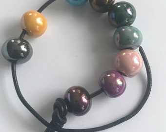 Puede incluir: Un brazalete de cuero negro con cuentas iridiscentes en tonos de azul, verde, marrón, rosa y amarillo.
