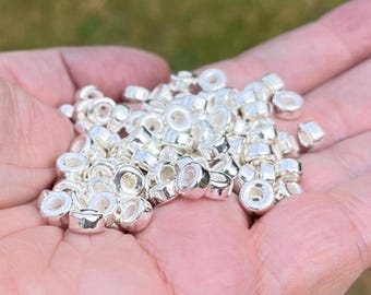 Puede incluir: Una colección de pequeñas cuentas redondas de color plata metálica con una superficie lisa y reflectante y un agujero central. Estas cuentas circulares tienen una forma uniforme y un acabado pulido, apareciendo como componentes individuales.