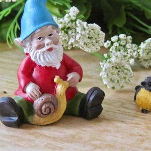 May include: A blue and white gnome figurine holding a yellow snail. The gnome is wearing a red shirt and green pants. A small yellow bird with black accents is sitting on the table.