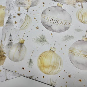May include: Wrapping paper featuring a repeating watercolor pattern of gold and grey Christmas ornaments, pine trees, and evergreen sprigs on a white background. The design includes small gold star accents and decorative lines on the baubles.