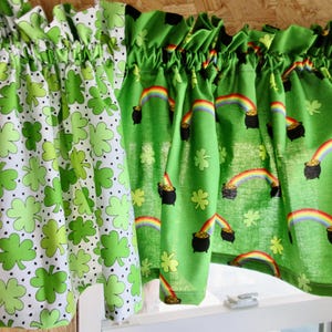 May include: Two green fabric valances with a shamrock and pot of gold design. The valance on the left has a white background with green shamrocks and black dots. The valance on the right has a green background with gold pots of gold, rainbows, and shamrocks.