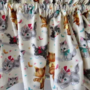 May include: Cream-colored fabric curtain with a repeating pattern of kittens, butterflies, and stars. The kittens are depicted in various colors, including gray, brown, and black, with red bows and teal bows. The butterflies and stars are in shades of pink and teal.