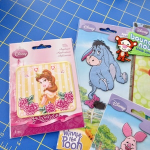 May include: Four packages of iron-on Disney character appliques. The appliques feature Belle from Beauty and the Beast, Eeyore from Winnie the Pooh, Tigger from Winnie the Pooh, and Piglet from Winnie the Pooh. The packages are pink, blue, green, and purple.