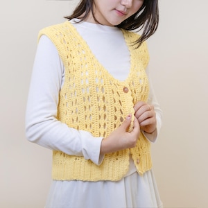 May include: A yellow crochet vest with a V-neck and two wooden buttons. The vest is worn over a white long-sleeved top. The crochet pattern has an open, airy design. The vest is a light, pastel yellow colour.
