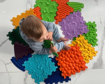 May include: A selection of brightly coloured, textured puzzle mats in a variety of shapes and shades, including orange, green, blue, and purple. The mats are arranged in a circular pattern. A baby is in the centre of the mats.