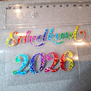 Könnte beinhalten: Ein regenbogenfarbener, glitzernder Aufbügler mit dem Wort Schulkind in Schreibschrift und der Zahl 2026 in fetter Schrift, platziert unter einem transparenten Kunststofflineal mit einer Länge von 15 cm als Größenvergleich.