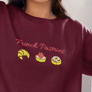 May include: Burgundy sweatshirt with the text "French Pastries" in a cursive font. Below the text are illustrations of a croissant, a strawberry tart, and a pastry. The sweatshirt is a solid color.