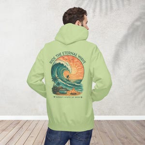 May include: A light green hoodie with a graphic on the back featuring a surfer riding a large wave at sunset. The text "RIDE THE ETERNAL WAVE" arches above the wave, and "SUNSET STATE OF MIND" is below. The hoodie has a hood and long sleeves.
