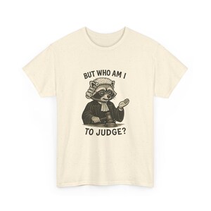 May include: Cream-colored t-shirt featuring a raccoon dressed as a judge, wearing a wig and holding a gavel. The text "BUT WHO AM I TO JUDGE?" is printed above and below the raccoon illustration. The shirt has a classic crew neck.