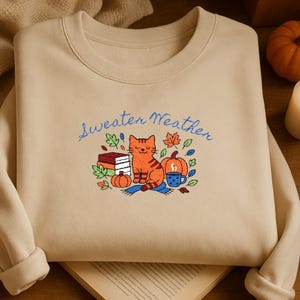 May include: Beige sweatshirt with the embroidered phrase "Sweater Weather" in blue script. The design features an orange tabby cat, pumpkins, books, and a mug of coffee, surrounded by fall leaves.