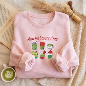 May include: A light pink sweatshirt with the text "Matcha Lovers Club" in red embroidery. Below the text are colorful embroidered images of matcha drinks. The sweatshirt is folded, with the sleeves visible.