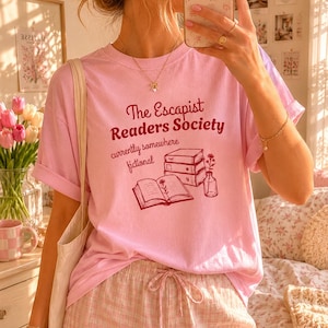 May include: A light pink t-shirt with the text "The Escapist Readers Society" in burgundy, along with illustrations of books and a vase of flowers. The shirt is paired with pink plaid shorts.