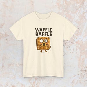 May include: Cream-colored t-shirt featuring the text "WAFFLE BAFFLE" above a cartoon waffle character with surprised eyes and open hands. The waffle has a brown color and is standing on two small feet.