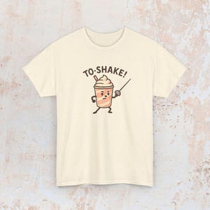 May include: Cream-colored t-shirt featuring a cartoon milkshake character with a fencing foil, and the text "TO-SHAKE!". The milkshake has a stern expression. The shirt is made of a soft material.