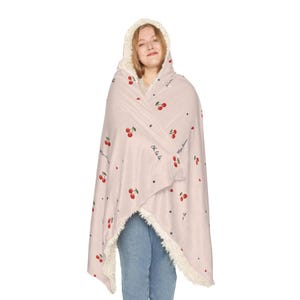 May include: A light pink hooded blanket with a cream-colored sherpa lining. The blanket is decorated with red cherry illustrations and small black stars. The words "Oh la la la" and "Mon amour" are written on the blanket.