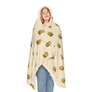 May include: A hooded blanket in a cream color with a repeating pattern of cartoon bees in yellow and black. The hood is lined with a fluffy, off-white material. The person is wearing blue jeans.
