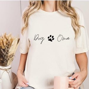 Könnte beinhalten: Cremefarbenes T-Shirt mit dem schwarzen Schriftzug "Dog Oma" und einem Pfotenabdruck. Die Person trägt zerrissene blaue Jeans und hält eine rosa Tasse. Im Hintergrund ein kleiner Tisch mit einer Vase mit Trockenblumen.